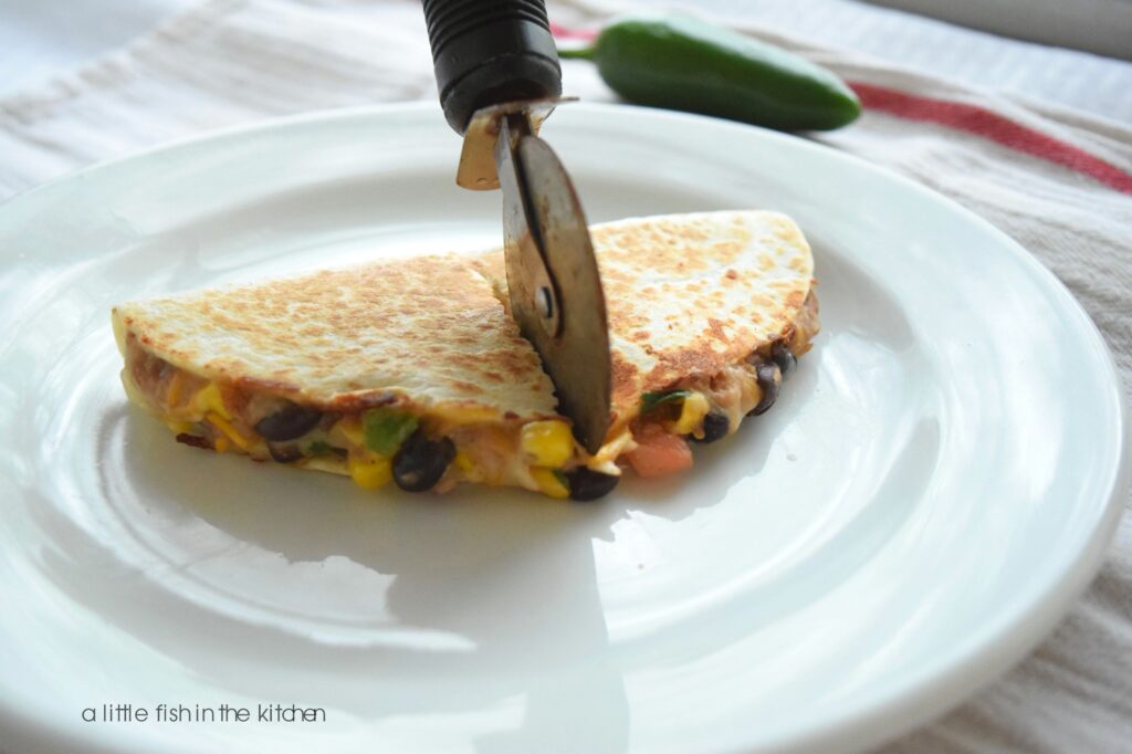 A single two-bean quesadilla is cut into two pieces with a pizza wheel, and is served on a white plate with a small red ramekin filled with red salsa. From the open cut sides, kernels of corn, whole black beans, refried beans, onions and cilantro are all visible in the quesadilla filling. 