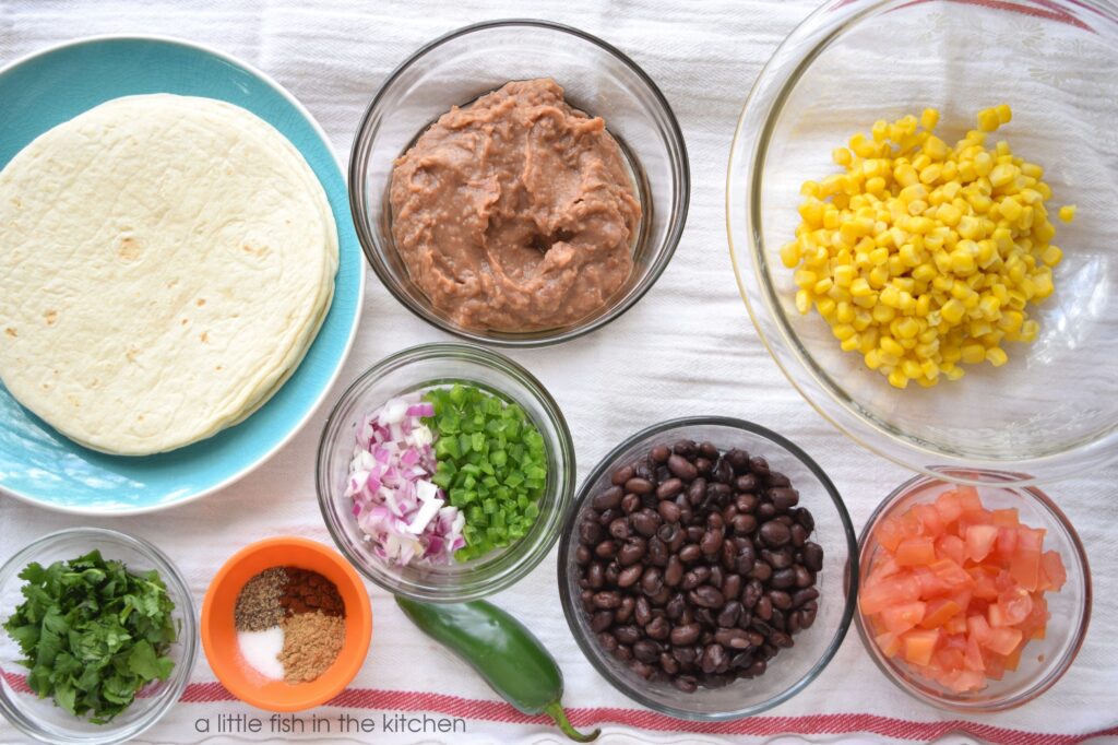 The ingredients for a spicy two bean southwestern quesadilla are gathered in separate prep bowls: yellow corn kernels, earthy refried beans, whole black beans, chopped tomato, finely diced red onion and peppers, taco seasoning and freshly torn cilantro are ready to layer into tortillas. 