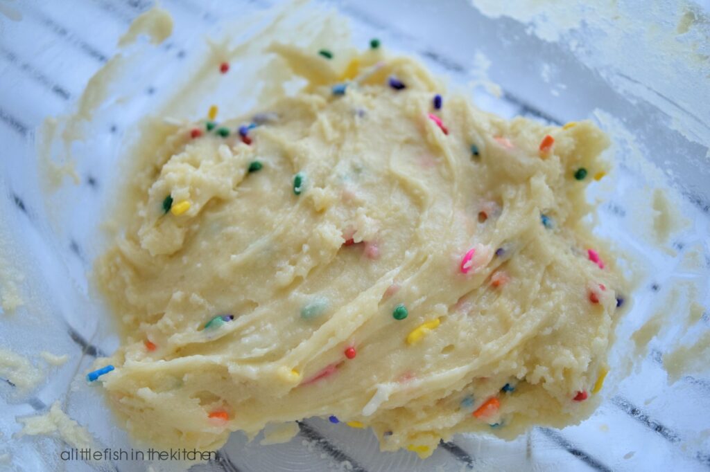 White cake mix cookie dough with rainbow sprinkles mixed in is ready to be portioned onto a baking sheet. The colorful sprinkles in every primary color stand out in the pale white cookie dough. 