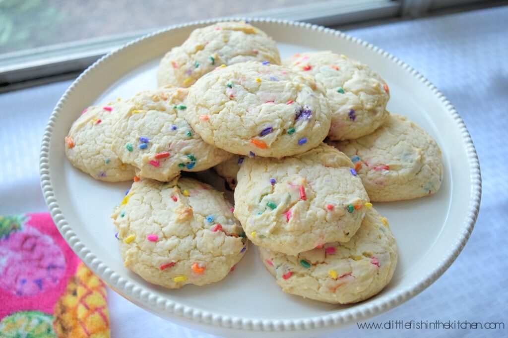 Confetti cake mix cookies are served on an ivory-colored ceramic cake stand with a pink tea towel laid out next to the cake stand. Colorful rainbowl sprinkles are strikingly visible in the soft white cake mix cookies. They are the perfect addition to a spring party dessert table. 
