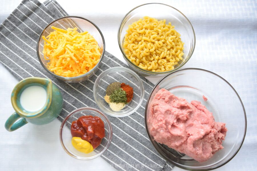 The ingredients for making skillet cheeseburger mac, uncooked macaroni noodles, shredded cheddar cheese, ground turkey meat, spices, ketchup, mustard and milk are gathered in separate bowls and ready to cook.