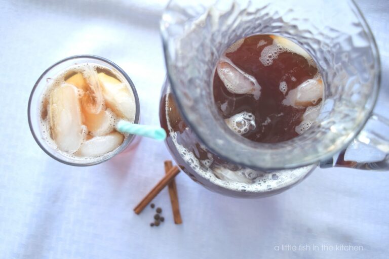 A clear glass pitcher filled with refreshing vanilla iced tea sits on a tabletop covered with a white linen tablecloth. A clear glass filled with iced tea and ice cubes is ready to enjoy. The glass has an teal and white striped plastic straw resting in it. 