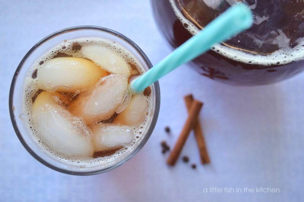  A freshly poured glass of iced tea has large iced cubes visible from the top and the glass is served with a teal and white striped reusable straw. A small pile of cinnamon sticks and whole all spices is slightly blurred beside the glass for decorative purpose.  