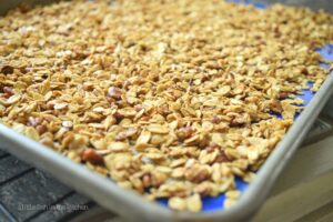 Golden, toasted granola is fresh from the oven and spread out on a large rimmed baking sheet. Honey-colored oats and toasted bits of pecan are visible from the top of the pan. 