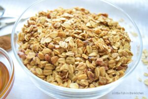 Freshmade honey-cinnamon granola is a clear bowl and ready to serve. Golden, crisp old-fashioned oats and toasted pecans pieces are visible throughout the bowl of homemade granola.  
