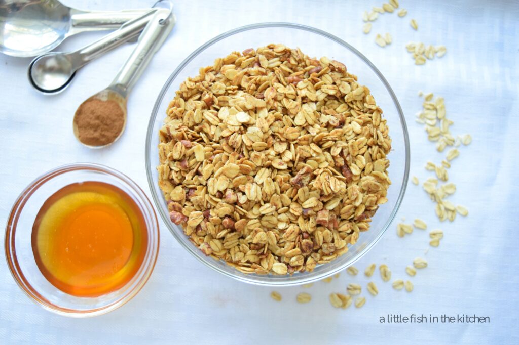 A clear glass bowl filled with homemade honey cinnamon granola is one a table covered with a white linen tablecloth. There are whole oats scattered around the bowl for decorative effect. There is a clear glass ramekin filled with golden-colored honey on the left side of the bowl as well as a spoon filled with ground cinnamon. Bits of toasted, chopped pecans and toasted old-fashioned oats coated with honey and cinnamon looked tempting in the serving bowl. 