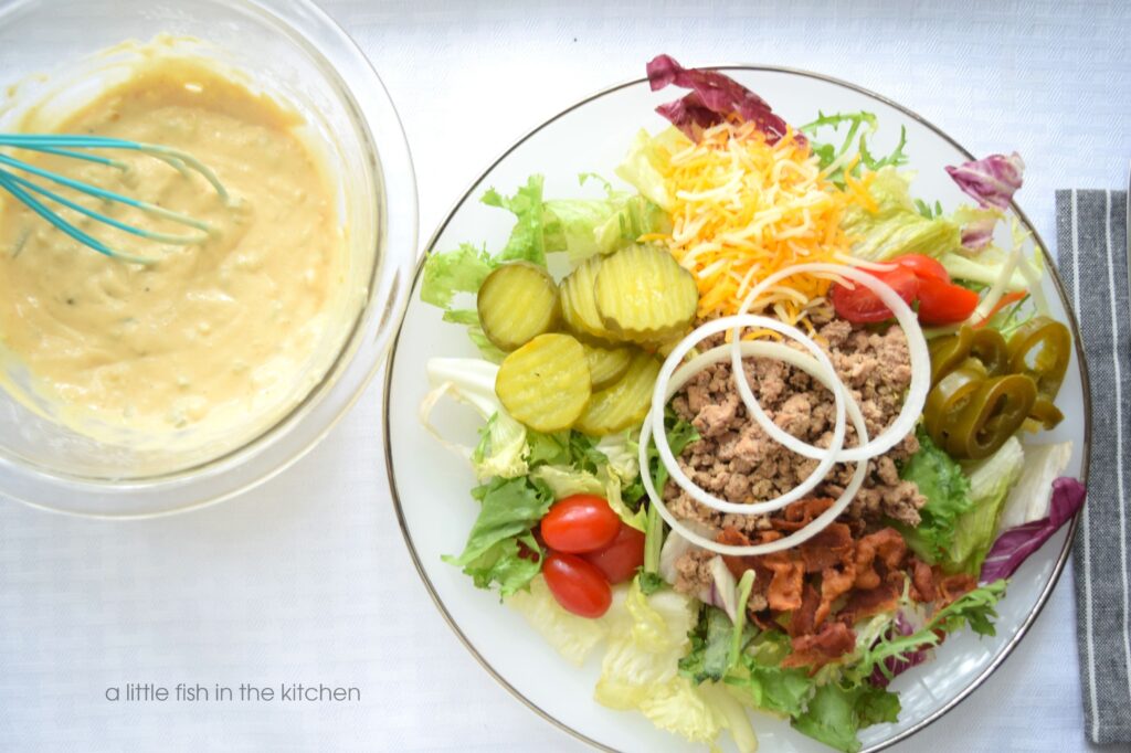 A loaded cheeseburger salad is on a white plate with a silver rim. A large portion of mixed greens is topped with sliced pickles, grated cheddar cheese, slice grape tomatoes, sliced pickled jalapenos, crumbled bacon and fresh slices of white onion. A small white bowl with homemade secret sauce salad dressing is right next to the plate. The salad dressing has a light coral hue and bits of diced pickles are visible in the creamy thousand island dressing. 