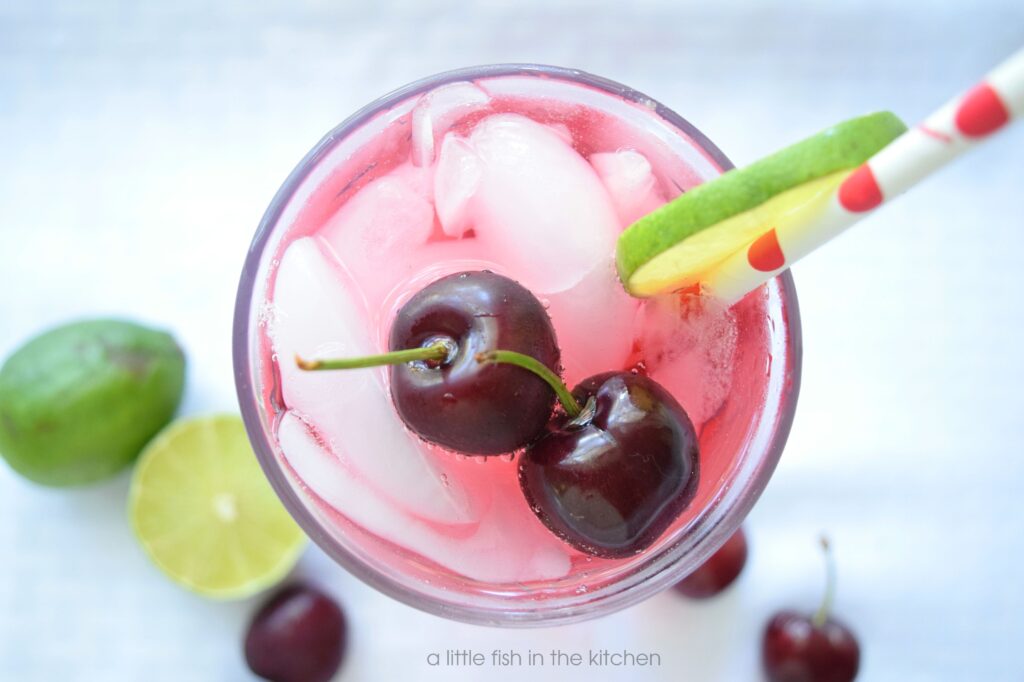 The cherry lime punch is garnished with two dark red whole cherries with stems and a slice of lime on the side of the glass. The garnishes rest invitingly on the top of the glass. 