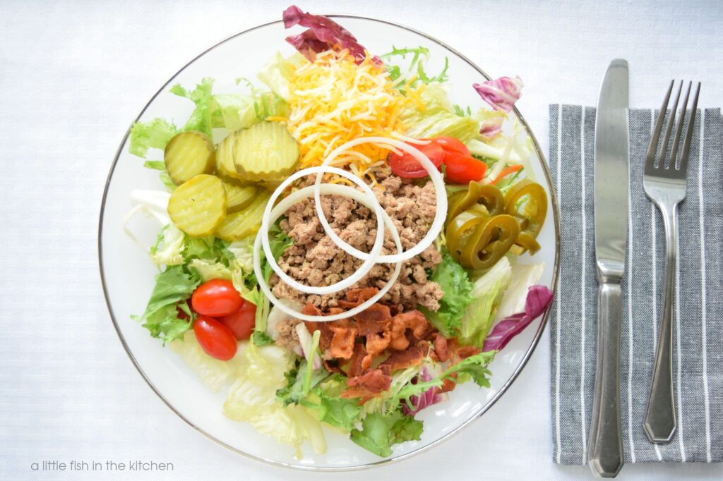 A loaded cheeseburger salad is on a white plate with a silver rim. A large portion of mixed greens is topped with sliced pickles, grated cheddar cheese, slice grape tomatoes, sliced pickled jalapenos, crumbled bacon and fresh slices of white onion. A gray napkin with white strips is next the plated with a fork and knife. This salad look satisfying and flavorful with all of it's hearty toppings. 
