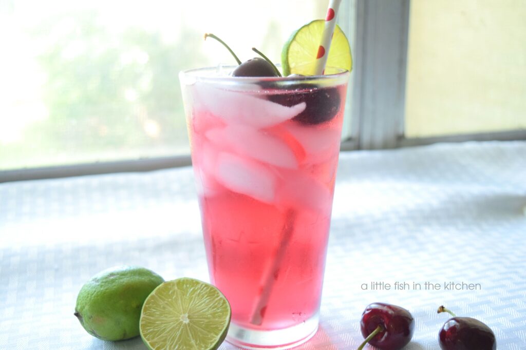A sparkling pink drink is served in a clear glass pint glass. There are crescent-shaped ice cubes visible in the glass as well as a white paper straw with red polka dots. The cherry lime punch is garnished with two dark red whole cherries with stems and a slice of lime on the side of the glass. 