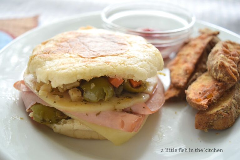 A hot, grilled mini muffaletta is served on a white plate with a serving a steak fries. melted cheese, slices of flavorful deli ham and delicious olive salad is visible between two slices of English muffin.