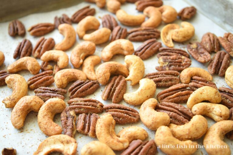 Roasted, seasoned pecan halves and whole cashews are cooling on a metal baking pan. The salt and seasonings are visible on the toasted nuts.