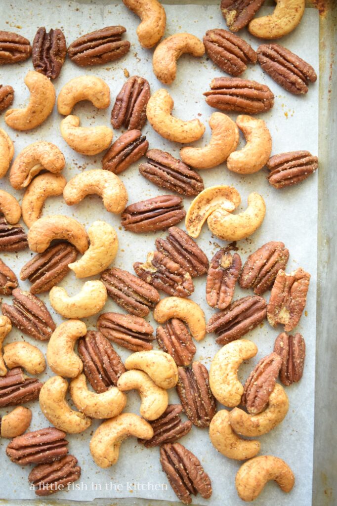 Roasted, seasoned pecan halves and whole cashews are cooling on a metal baking pan. The salt and seasonings are temptingly visible on the toasted nuts.