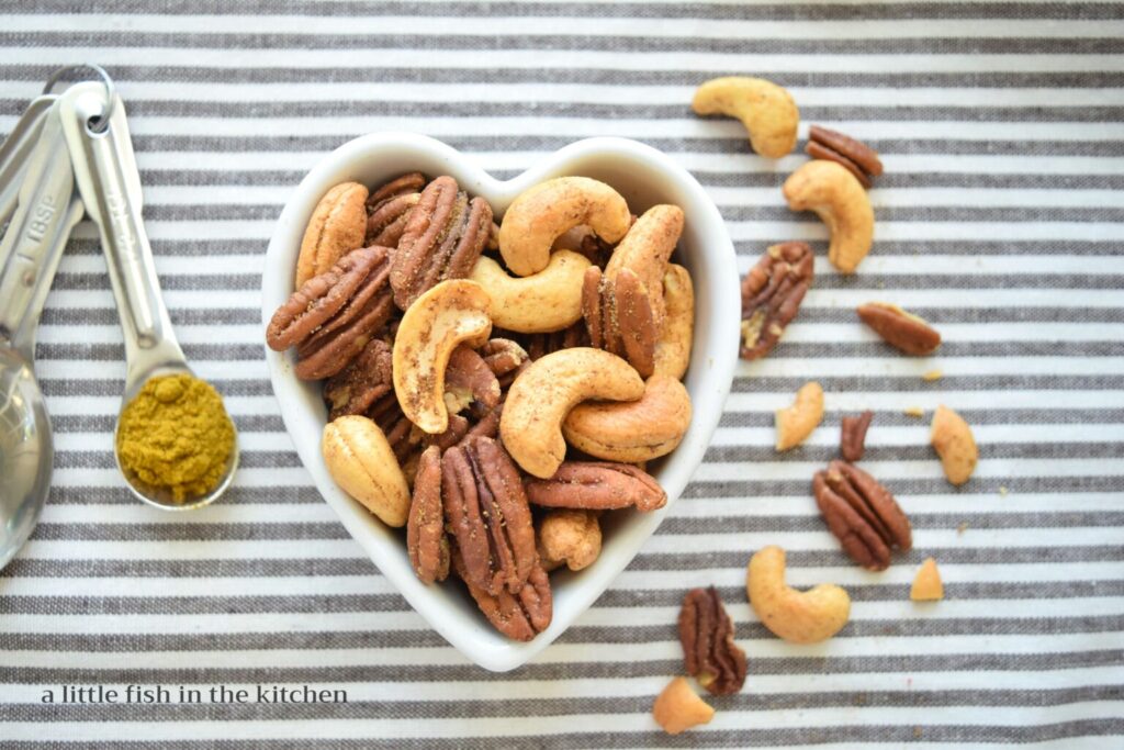 Spicy curry mixed nuts are served in a cute heart-shaped ceramic ramekin.  Roasted, seasoned pecan halves and whole cashews are featured in this recipe. The white ramekin is sitting a gray and white striped tea towel with a few nuts and a teapoons filled with curry powder just beside it for a decorative effect in the photo. 
