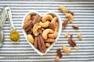 Spicy Curry seasoned nut mix is presented in small, white, heart-shaped bowl. The bowl is laying on a gray and white striped tea towel. A measuring spoon filled with golden curry powder and a small array of whole nuts lay beside the bowl for a pretty presentation. 