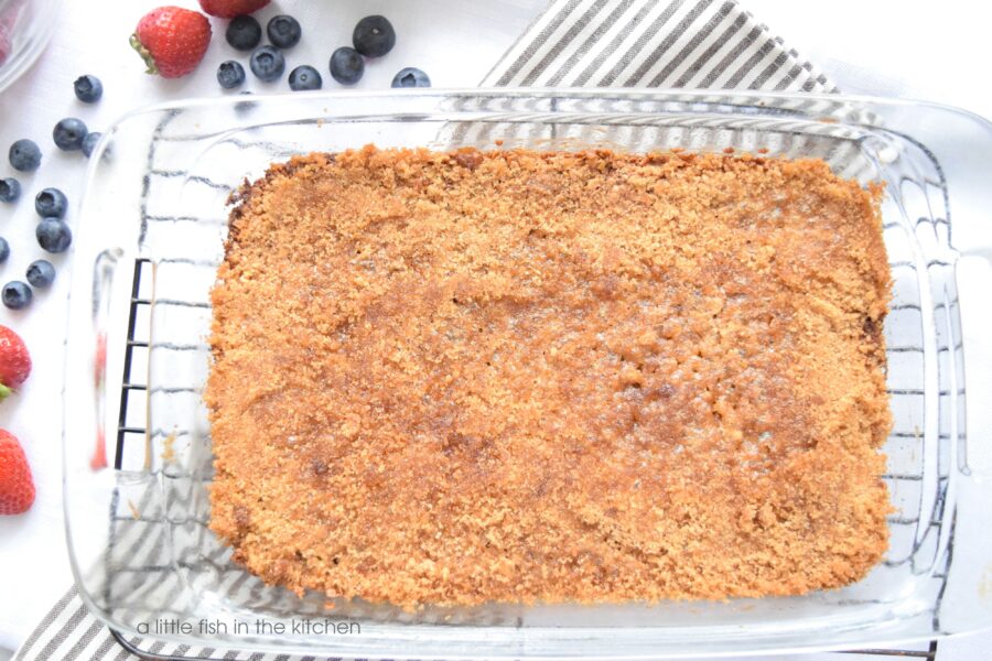 A freshly baked pan of brown sugar baked oatmeal is cooling on a metal rack. The oatmeal is baked in a clear glass 9x13 inch dish, and a gray and white striped tea towel is beneath the cooling rack, it's visible through the glass baking dish. A few fresh berries are scattered along the left side of the baking dish for a decorative effect. 