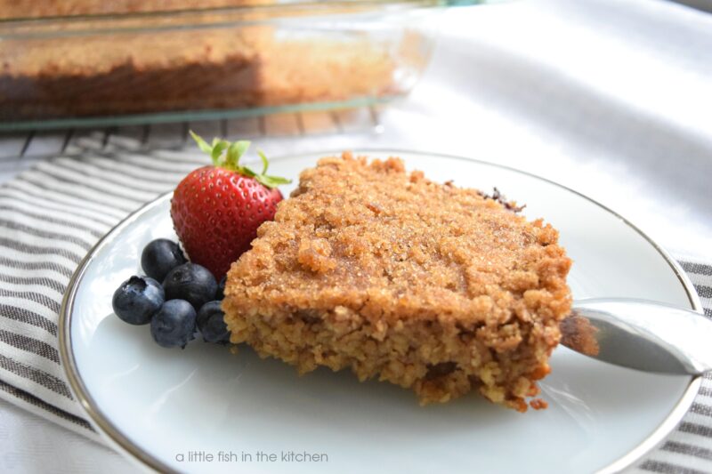 A single slice of Spiced brown sugar baked oats is on a small white plate with a silver rim. The crispy brown sugar coating on top of the oats looks tempting. A small portion of fresh strawberries and blueberries is served beside a slice of warm, baked oatmeal. 