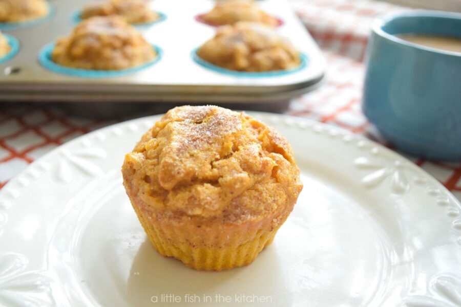 A single cinnamon and sugar dusted pumpkin apple muffin sits on a white ceramic dessert place. A light blue mug filled with coffee sits just behind the plate. The muffin tin with the remainder of the warm pumpkin muffins is behind the plate and slightly blurred in the picture. 