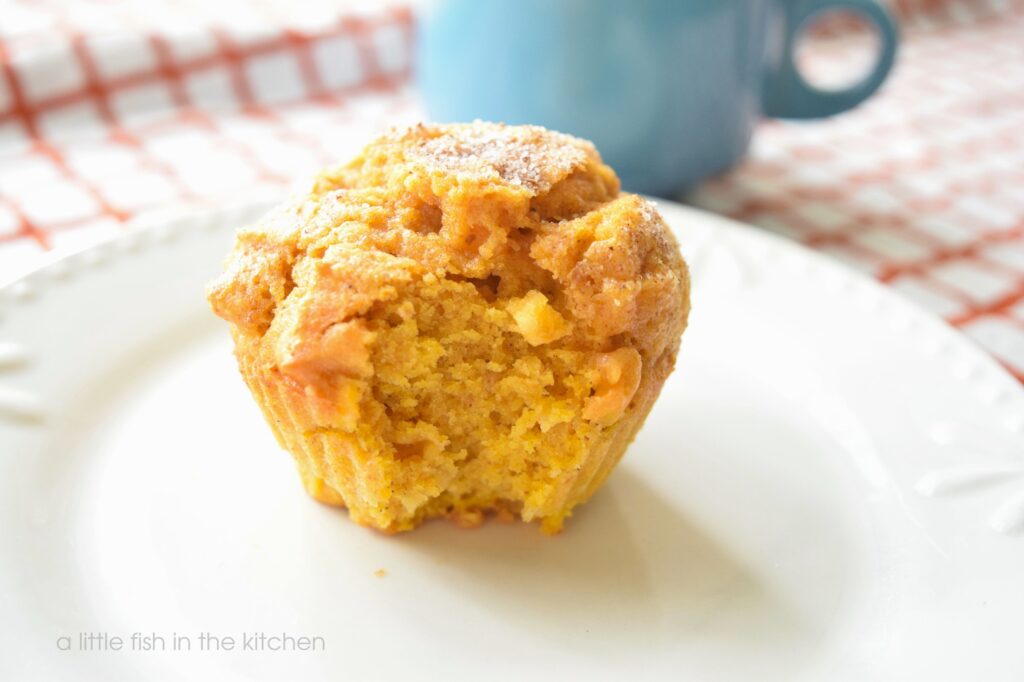 A single made-from-scratch pumkin apple muffin is served on a small, white ceramic plate. A bite of been taken from the muffin exposing the tender, amber-colore crumb as well as bits of fresh apple baked into the spiced pumpkin muffins. The plate sits on a white tea towel with orange stripes and a light blue coffee mug is slightly blurred in the background. 
