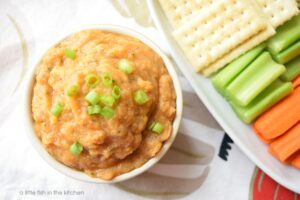 The top of a small bowl filled with amber-colored pumpkin dip is garnished with sliced green onions. A small rectangular white plate sits beside the bowl with saltine crackers and fesh celery and carrot sticks, ready for dipping. 