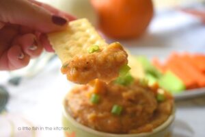 A generous dollop of amber-colored pumpkin dip is on the edge of a saltine cracker. The dip is smooth and thick and looks inviting. The small bowl of dip and the plate of crackers and vegetable sticks are slightly blurred in the background. 