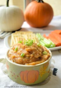 A small, light green decorative bowl is filled with amber-colored pumpkin-bean dip! It's garnishes with sliced green onion sprinkled over the top. A small plate with saltine crackers and fresh veggies sits behind the bowl of dip. Two small whole, fresh pumpkins, one white, one orange are slightly blurred in the background. 