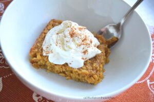 A single square slice of Pumpkin Pie Baked Oatmeal is resting a white bowl, it is topped with a swirl of whipped cream and a sprinkle of cinnamon. The bowl sits on top of a burnt orange colored place mat with floral pattern. A spoon is resting inside the bowl. 