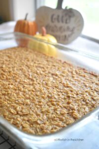 A clear baking dish is shown from the corner and a fresh batch of Pumpkin Pie Baked Oatmeal is shown from the top. Oats are seen baked into the top of pumpkin filling. 3 decorative small pumpkins are slightly blurred in the background. 