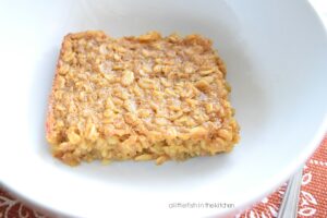 A single square slice of Pumpkin Pie Baked Oatmeal is resting a white bowl. The bowl sits on top of a burnt orange colored place mat with floral pattern. A spoon is resting beside the bowl. 