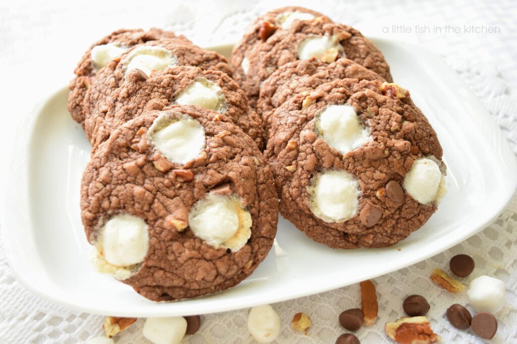 Six chocolatey Mississippi Mud cookies are arranged in two rows on a small white ceramic platter. Three melty marshmallow, chocolate chips and bits of toasted pecans are visible on the tops of the cookies. They are look so rich and delicious. They ceramic platter sits on a white lace doily and a few small marshmallows, chopped pecans and chocolate chips are scattered around the platter for a decorative effect.