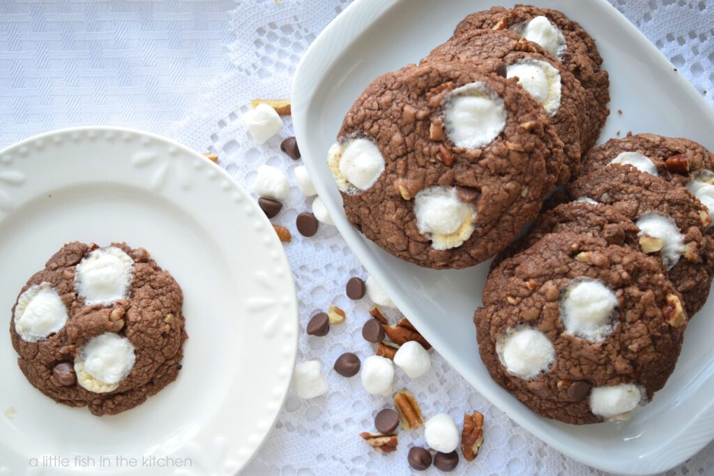 Six rich-looking chocolate cookies are arranged on a small white platter and a single cookie is next to the platter with a single Mississippi Mud cookie on top. The plate and platter on served atop a dainty, white, lace doily for a lovely rustic effect.