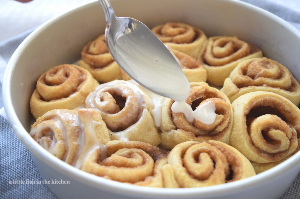 Fresh baked cinnamon rolls are in round metal baking dish. A spoon is hovering above the pan of cinnamon rolls and is drizzling white glaze over the rolls. 