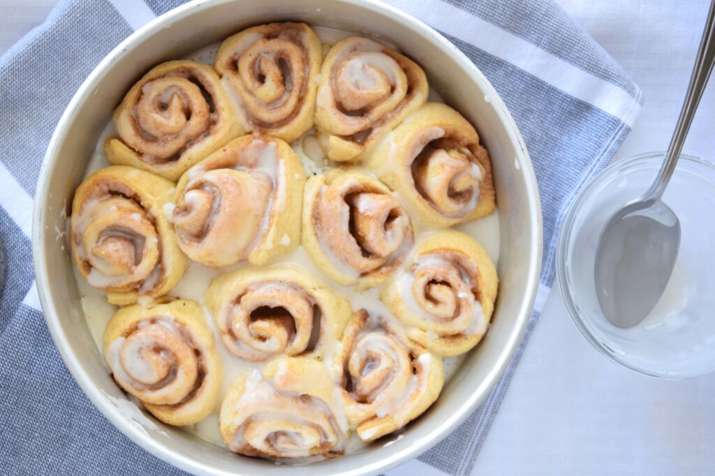 Freshly iced cinnamon rolls are in a round metal baking dish and are ready to serve. The pan sits on a gray and white striped tea towel. A small clear glass bowl with the remainder of the white glaze and a spoon sit beside the baking pan. 