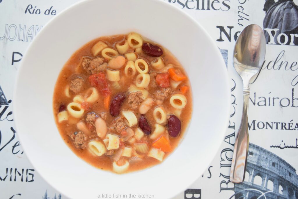 An amber-colored brothy soup is in a white bowl, ready to serve. A metallic silver spoon rests beside the bowl. Bits of ground meat, whole white and dark kidney beans, carrots, chunks of tomato, diced onions, are all visible inside the bowl of pasta classic fagioli soup.