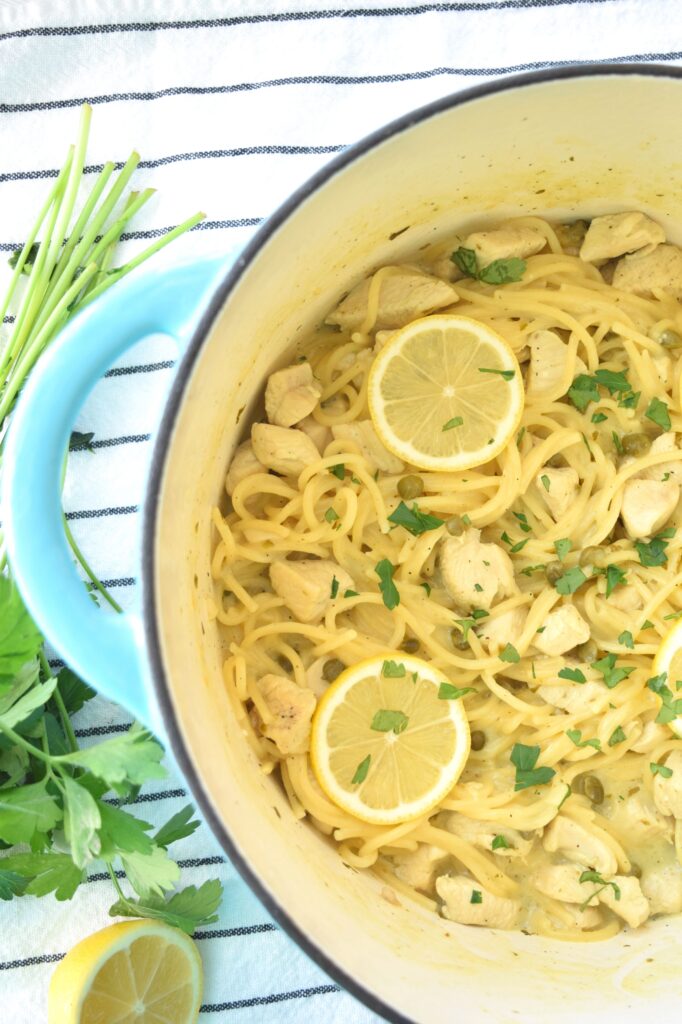 One half of a teal Dutch oven is seen and in it cooked spaghetti, bits of chicken and capers are visible. Lemon slices and chopped parsley are scatter on top of the pasta for garnish. A bunch of fresh parsely is visible on the side of the pot. 