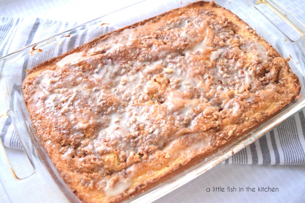 A Honey Bun Cake is fresh out of the oven and cooling on a small rack. A gray and white tea towel is underneath the clear glass baking dish! 