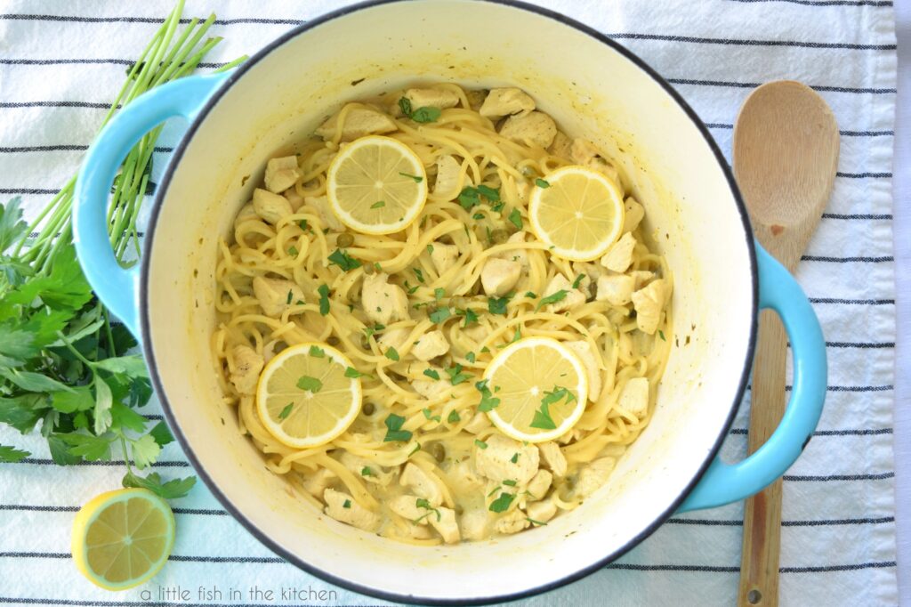 Cooked spaghetti, bits of chicken, are resting in a teal colored Dutch oven. The pasta is garnished with chopped parsley and lemon slices. A wooden spoon and a bunch of fresh parsely rest beside the pot on top of a white towel with think black stripes. 