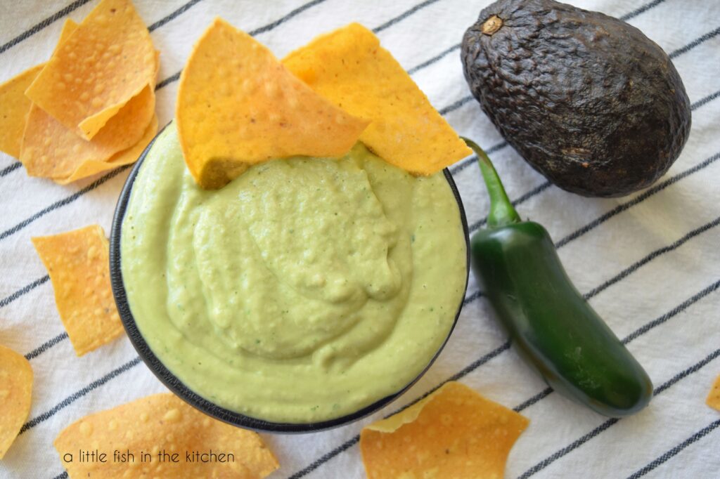 Creamy Avocado Salsa in a small black serving bowl.  A jalapeño pepper, scattered tortilla chips and a whole avocado are next to the bowl. 