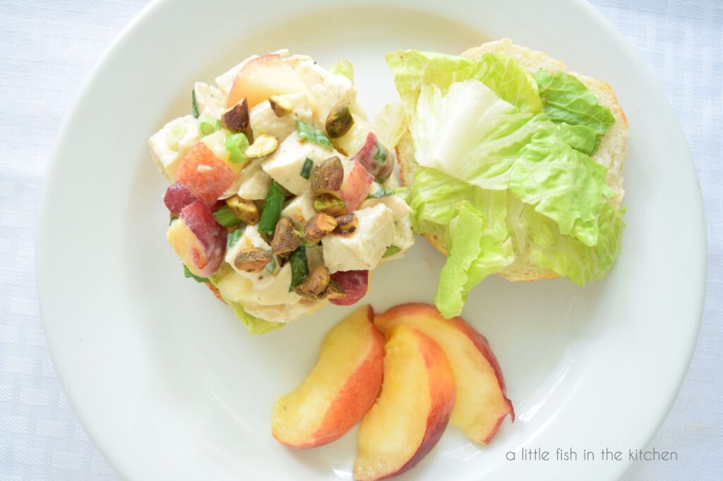 Both sides of a bagel are shown on a white plate. One side is topped with creamy chicken salad and other is topped with a lettuce. A portion of sliced fresh peach sits on the plate for garnish. 