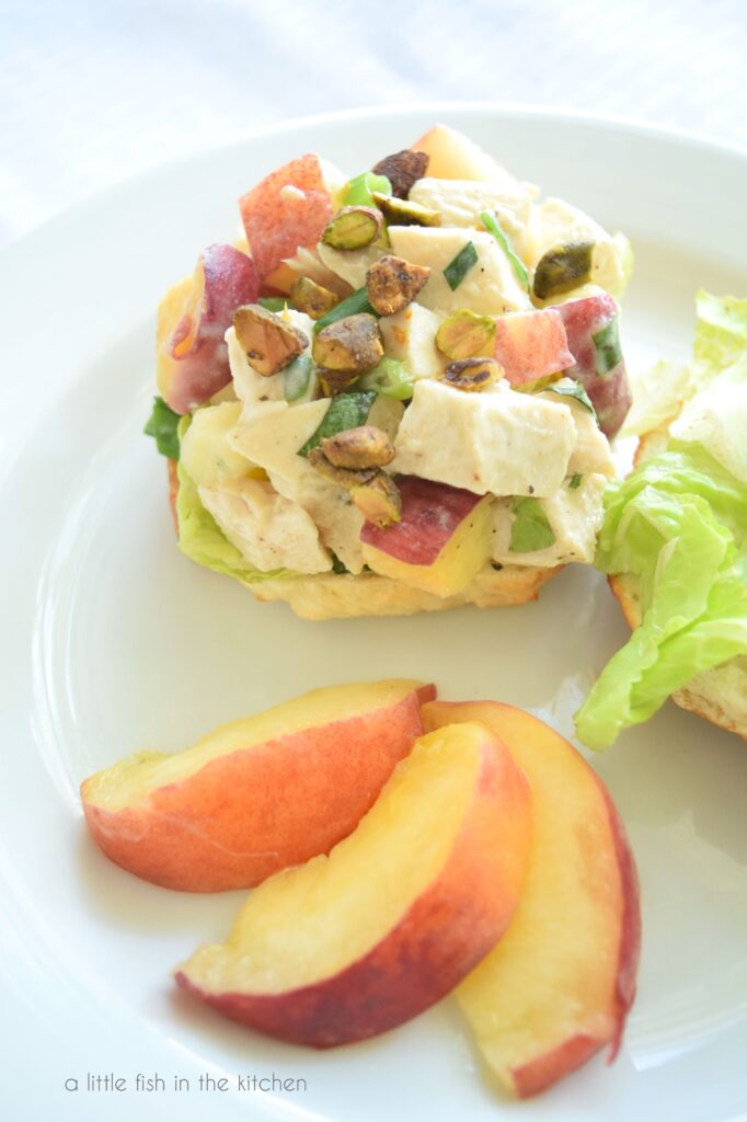 Colorful, creamy chicken Salad sit atop a bagel on a white plate. A portion of a sliced, fresh peach is beside the sandwich. Bits of diced chicken, fresh peaches, green onion and lettice are visible in the filling. 