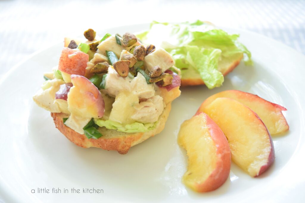 Colorful Peach-Pistachio Chicken Salad sit atop a bagel on a white plate. A portion of a sliced, fresh peach is beside the sandwich. Bits of diced chicken, fresh peaches, green onion and lettice are visible in the filling. 