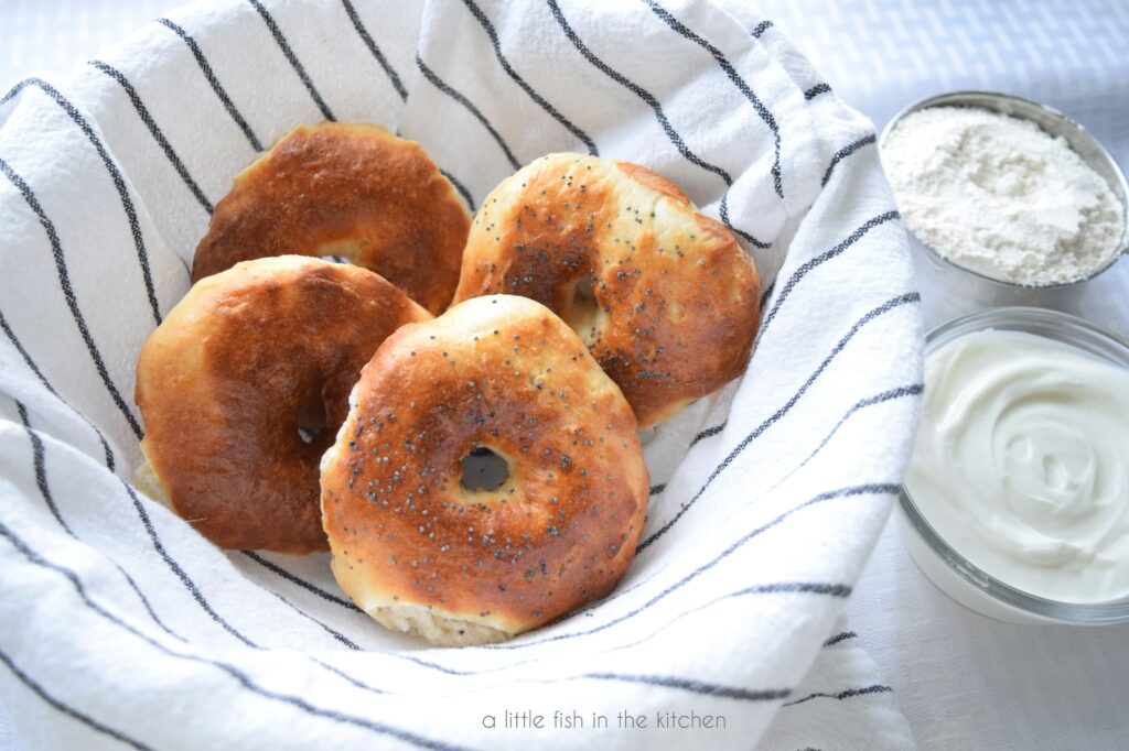 Fresh two-ingredient bagels shown with flour and yogurt beside basket. The tops are perfectly golden brown and poppy seeds are on the tops of two bagels. 