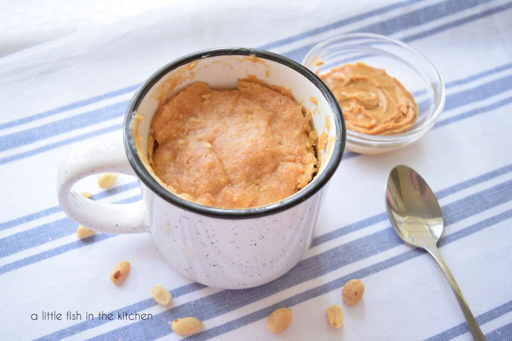 A mug with peanut butter cake inside sits on a blue and white table runner. A small bowl with peanut butter and a spoon sits beside it.