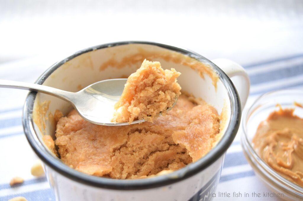 A spoon sits on top of the peanut butter mug cake in the mug. It has a bite of moist peanut butter cake in it. 