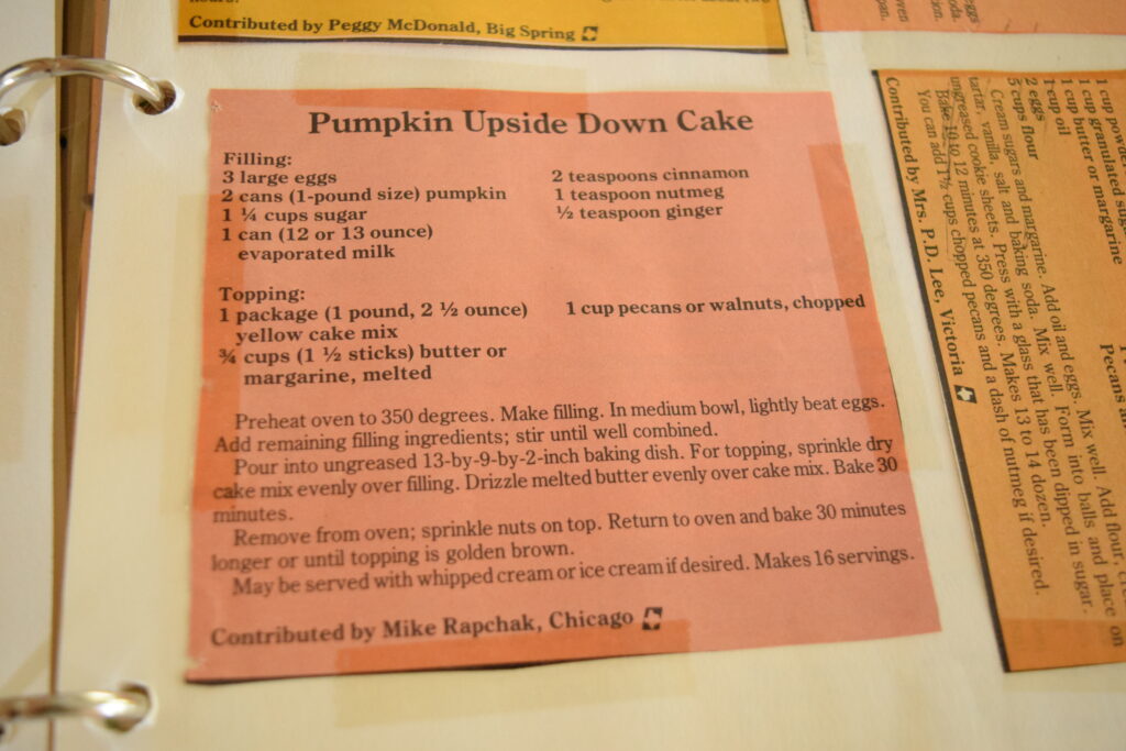A photo of the original old recipe clipping with the title "Pumpkin Upside Down Cake . The recipe clipping is made of light red paper and it's taped to a white cardbord folder divider. The ring of the spiral notebook is visible. 