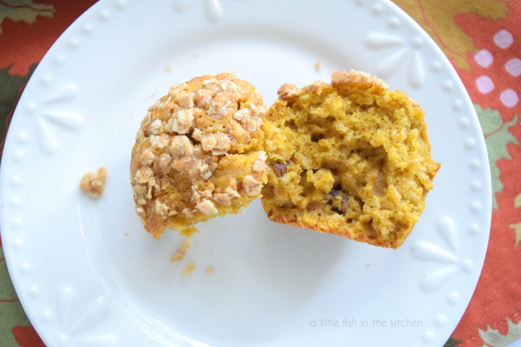 A single pumpkin oat streusel muffins  torn in half on a plate, pecan pieces are visible in the orange-tinted crumb of the muffin.