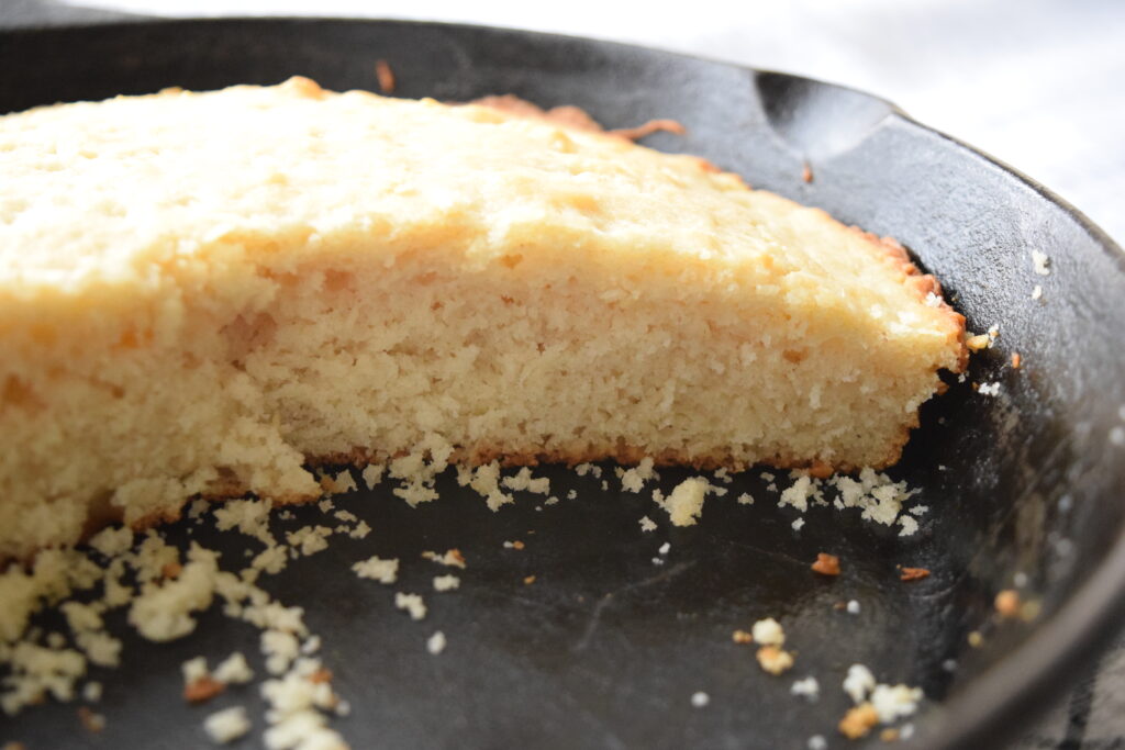 A round loaf of golden, soft skillet biscuit bread is cut in half as it sits in a black cast iron skillet. The golden, fluffy texture of the bread is shown from the inside. 