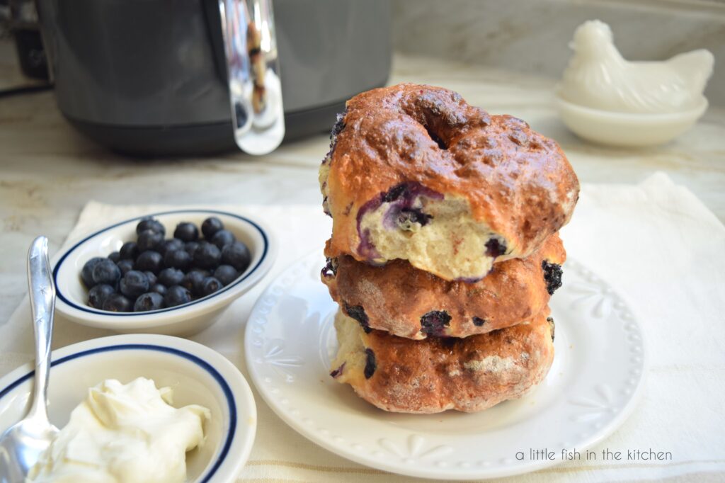 A stack of three blueberry bagels sits on top of a small white plate. Two small bowls are beside the plate. One bowl has cream cheese and a spoon and the other bowl holds fresh blueberries. An air fryer and a decorative milk glass rooster are slight blurred in the back ground. 