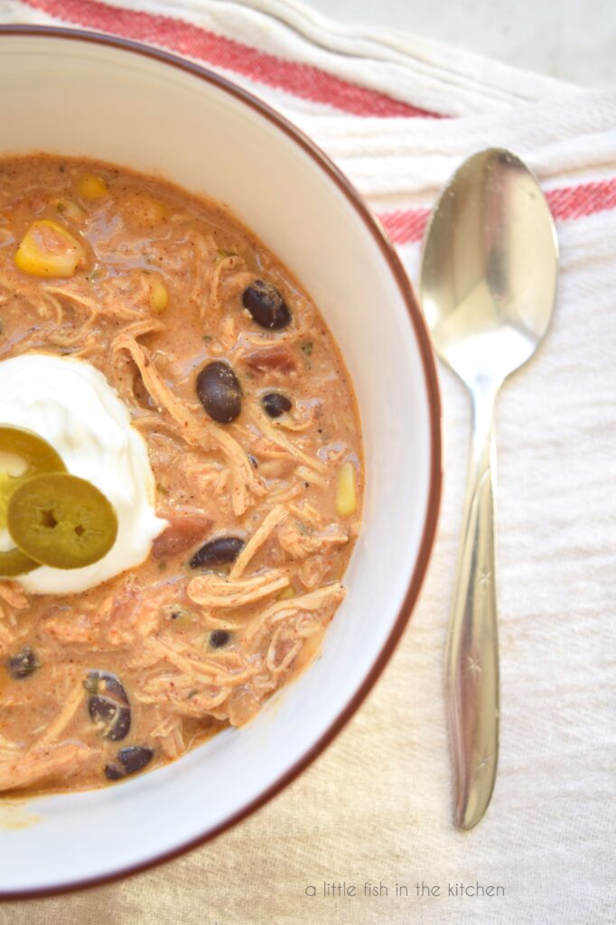 A bowl filled with auburn colored cream cheese chicken chili sits inside a white bowl with a brown rim. The bowl is on a white towel with red trim. Corn, black beans and bits of tomato are visible in the soup. A spoon lays beside the bowl. 