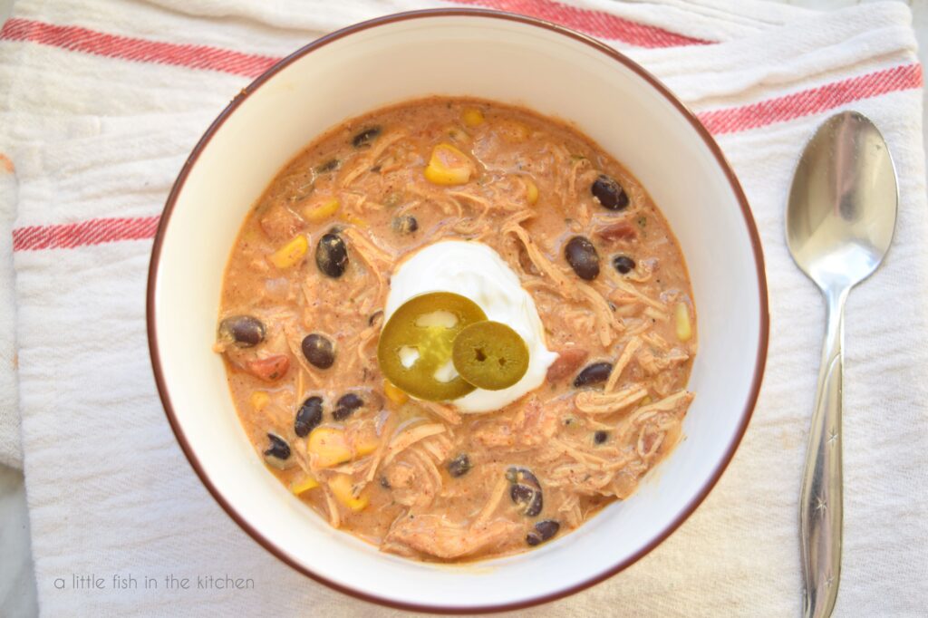 A bowl filled with auburn colored cream cheese chicken chili sits inside a white bowl with a brown rim. Corn, black beans and bits of tomato are visible in the soup. A spoon lays beside the bowl. 